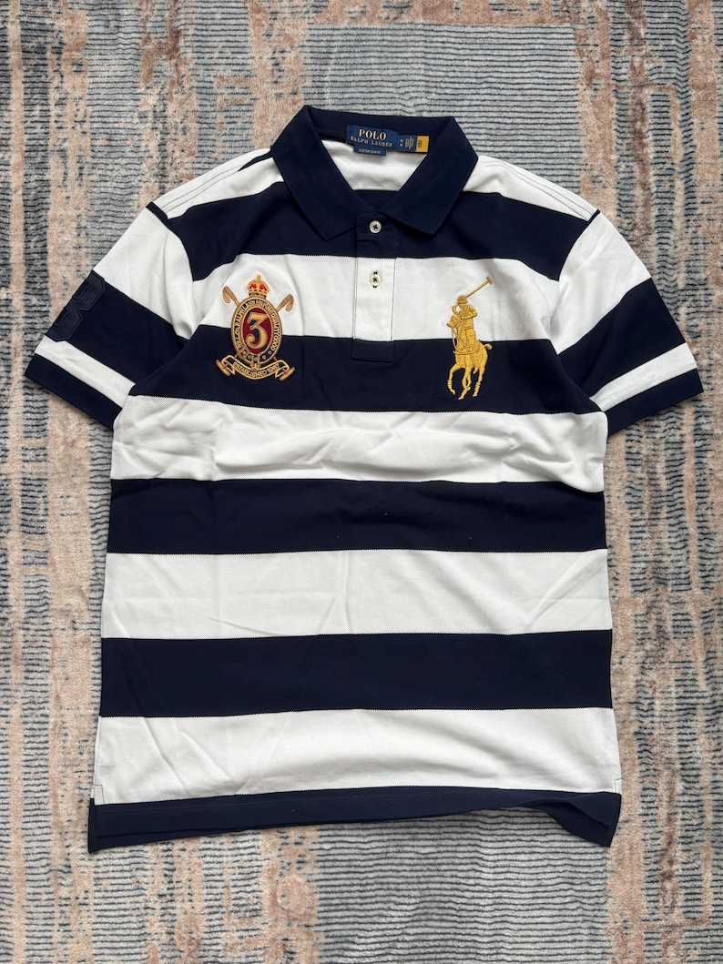 Puede incluir: Polo a rayas azul marino y blanco con mangas cortas. La camisa tiene cuello azul marino y tapeta de tres botones. Cuenta con un logotipo ecuestre dorado y un escudo con el n&uacute;mero 6 bordado en el pecho.