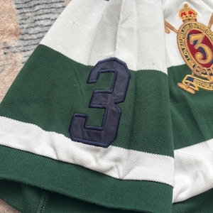 Puede incluir: Polo a rayas verdes y blancas con un parche azul marino con el n&uacute;mero 3 en la manga. La camisa presenta un escudo con bordados dorados y el n&uacute;mero 3.