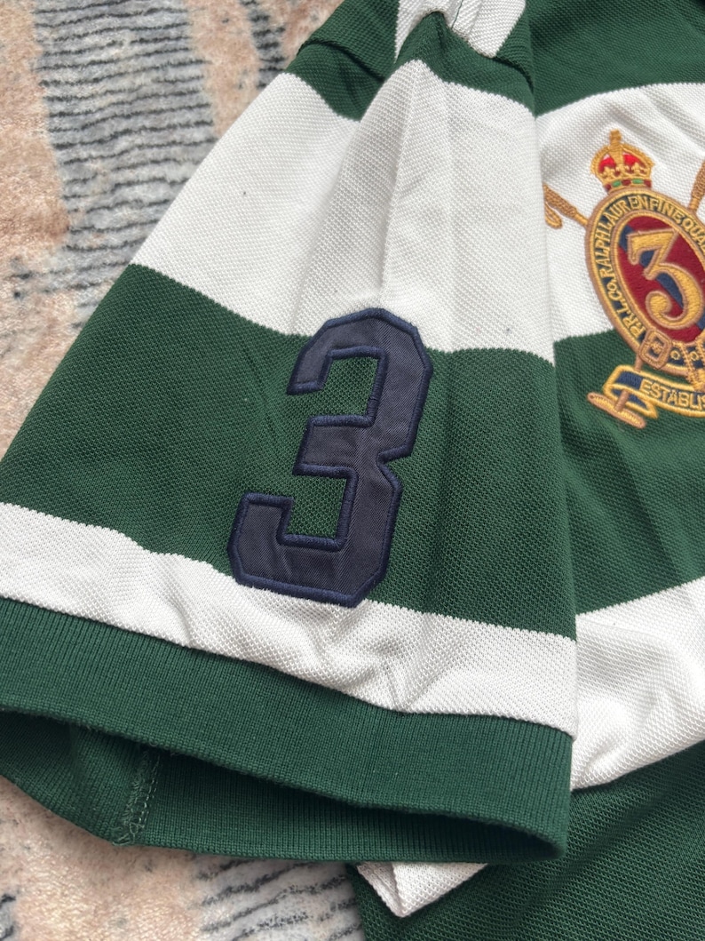 Puede incluir: Polo a rayas verdes y blancas con un parche azul marino con el n&uacute;mero 3 en la manga. La camisa presenta un escudo con bordados dorados y el n&uacute;mero 3.