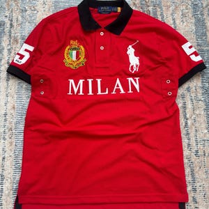May include: A red polo shirt with a black collar and sleeves. The shirt features the word "MILAN" in white block letters, a white polo player logo, and the number "5" on the sleeves. A crest is also visible.