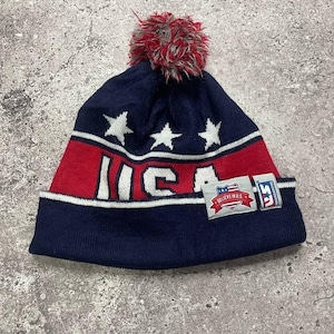 Op de afbeelding: Blauwe en rode gebreide beanie met een witte pompon bovenop. De beanie heeft witte sterren en het woord "USA" in witte letters op een rode streep. De beanie heeft een label met de tekst "Believe in US" met een kleine Amerikaanse vlag.