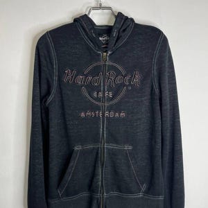 Könnte beinhalten: Ein schwarzer Kapuzenpullover mit Reißverschluss und dem Hard Rock Cafe-Logo in einem kreisförmigen Design. Das Logo und das Wort "AMSTERDAM" sind in einer helleren Farbe gestickt. Der Hoodie hat eine Vordertasche und hängt an einem Metallbügel.