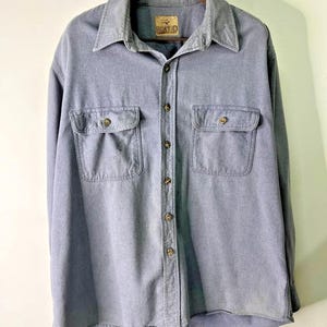 Puede incluir: Una camisa azul abotonada con dos bolsillos en el pecho y un cuello. La camisa tiene un aspecto descolorido y una pequeña etiqueta que dice "REDHEAD". La camisa está colgada de una percha de metal.