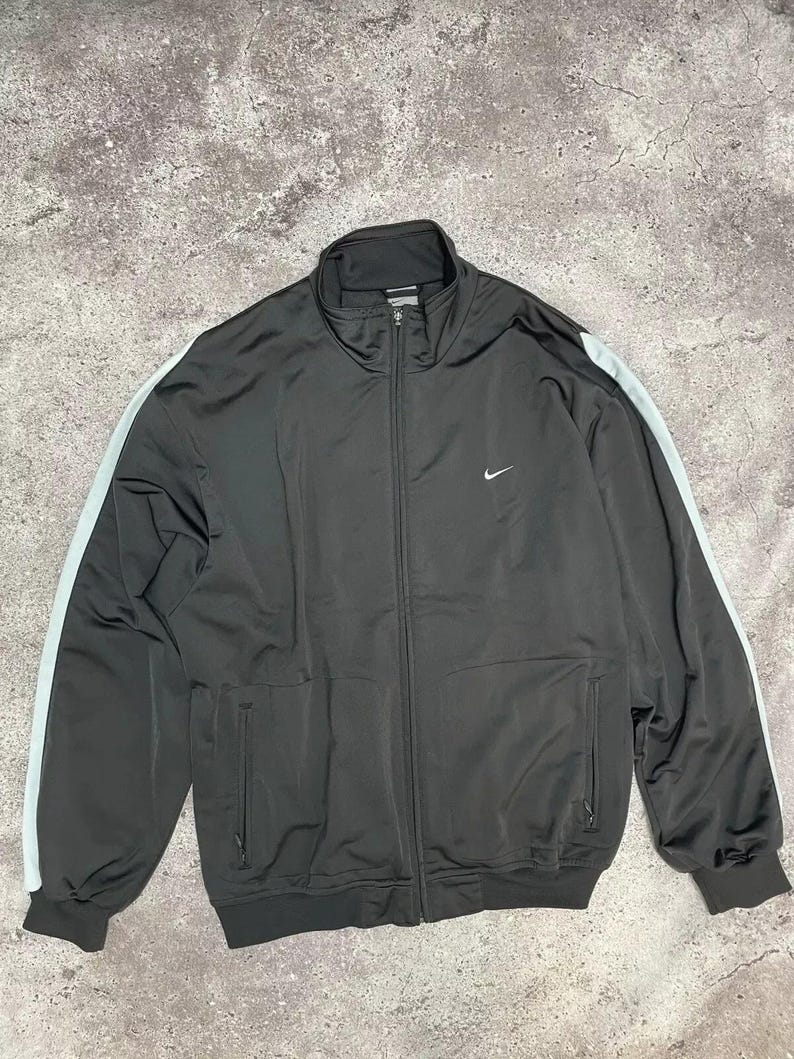 Puede incluir: Chaqueta deportiva negra con cremallera completa, dos bolsillos delanteros y un peque&ntilde;o logotipo blanco de Nike en el pecho. La chaqueta tiene rayas azul claro en las mangas.