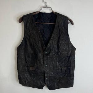 Könnte beinhalten: Eine braune Lederweste mit Knopfleiste und mehreren Taschen. Die Weste hat ein gestepptes Design und hängt an einem Kleiderbügel.