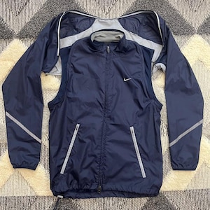 Puede incluir: Una chaqueta cortavientos azul marino con cierre de cremallera y un logotipo blanco de Nike en el pecho. La chaqueta tiene un forro de malla y detalles reflectantes en las mangas y los bolsillos.