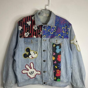 Pode incluir: Uma jaqueta de ganga azul claro com patches coloridos com o Mickey Mouse. Os patches incluem o rosto do Mickey Mouse, sua icônica luva branca e um padrão repetido de cabeças do Mickey Mouse.