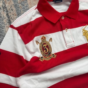 Puede incluir: Polo a rayas rojas y blancas con cuello cl&aacute;sico. La camisa presenta un emblema dorado con el n&uacute;mero 3 y el texto "ESTABLISHED 1967". La marca "POLO RALPH LAUREN" es visible en el cuello.
