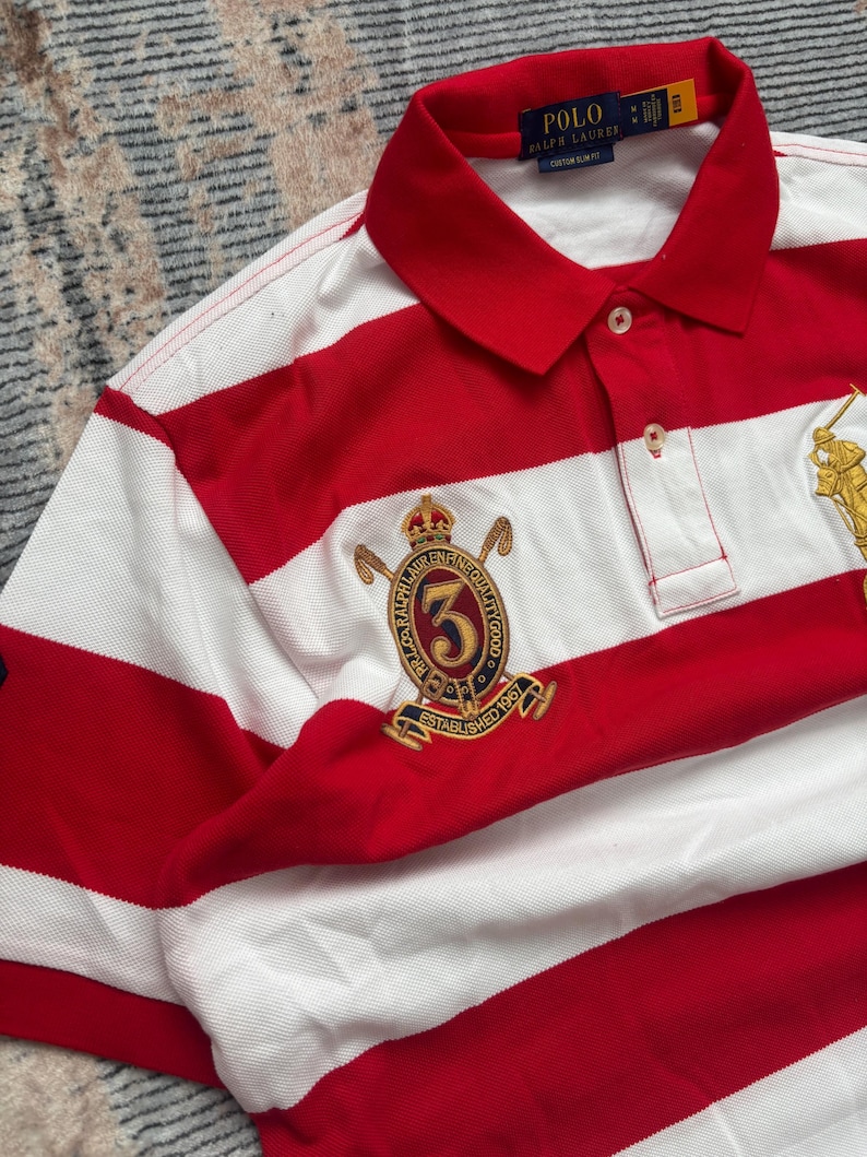 Puede incluir: Polo a rayas rojas y blancas con cuello cl&aacute;sico. La camisa presenta un emblema dorado con el n&uacute;mero 3 y el texto "ESTABLISHED 1967". La marca "POLO RALPH LAUREN" es visible en el cuello.