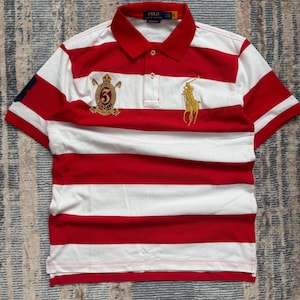 Puede incluir: Polo a rayas rojas y blancas con mangas cortas. Presenta un logotipo dorado bordado de un jinete a caballo y un escudo con el n&uacute;mero 5. La camisa tiene un cuello cl&aacute;sico y una tapeta de tres botones.