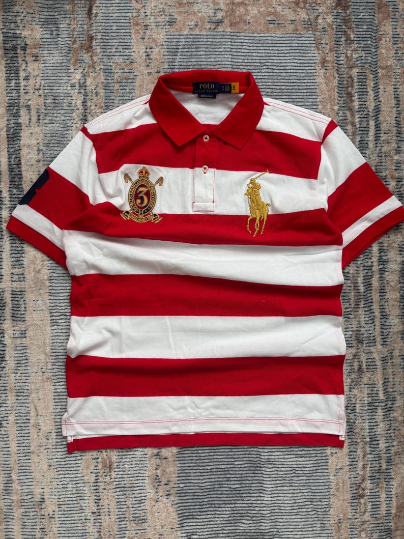 Puede incluir: Polo a rayas rojas y blancas con mangas cortas. Presenta un logotipo dorado bordado de un jinete a caballo y un escudo con el n&uacute;mero 5. La camisa tiene un cuello cl&aacute;sico y una tapeta de tres botones.