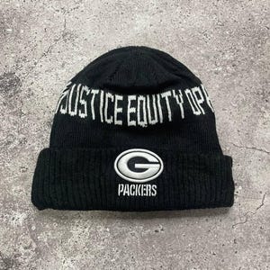 Puede incluir: Gorro de punto negro con una inscripción blanca "Justice Equity Opportunity" y un logotipo verde y blanco de los Green Bay Packers con el texto "Packers".