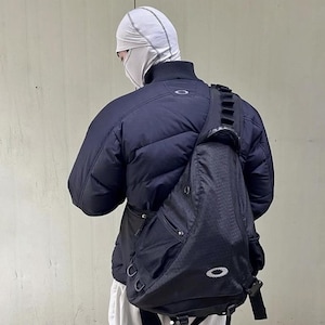 May include: A black sling bag with a textured pattern and the Oakley logo. The bag is worn over the shoulder and across the chest. The person is wearing a black puffer jacket and a white balaclava.