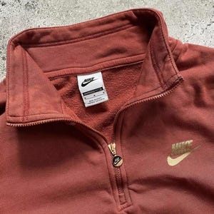 May include: A rust-colored Nike quarter-zip sweatshirt with a gold Nike logo on the right side and a white Nike tag on the left side. The tag reads "Oversized Fit L/G" and "Made in Egypt."