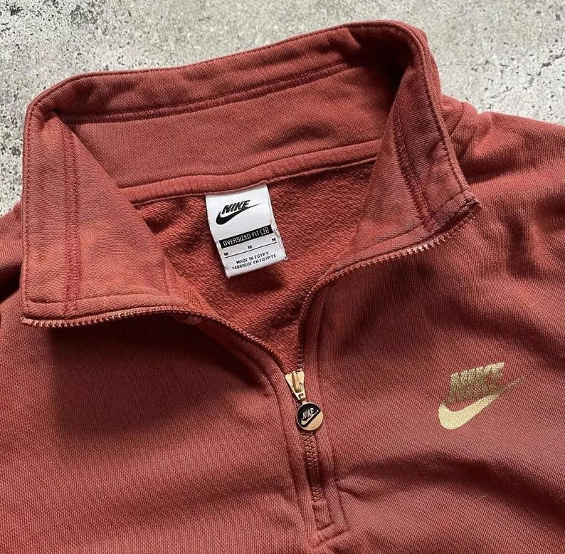 May include: A rust-colored Nike quarter-zip sweatshirt with a gold Nike logo on the right side and a white Nike tag on the left side. The tag reads "Oversized Fit L/G" and "Made in Egypt."