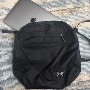 May include: A black, lightweight backpack with a zippered top and a small white logo on the lower right. A silver laptop is partially visible inside the bag. The bag has a drawstring closure and a shoulder strap.