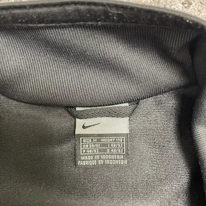 Puede incluir: Primer plano de una chaqueta negra con una etiqueta. La etiqueta incluye el logotipo de Nike e informaci&oacute;n de tallas: TALLA M, ALTURA 178, GB 39/41, F 48/52 y D 48/52. La etiqueta tambi&eacute;n indica "MADE IN INDONESIA".