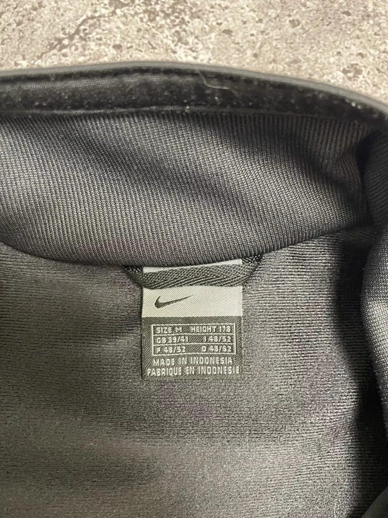 Puede incluir: Primer plano de una chaqueta negra con una etiqueta. La etiqueta incluye el logotipo de Nike e informaci&oacute;n de tallas: TALLA M, ALTURA 178, GB 39/41, F 48/52 y D 48/52. La etiqueta tambi&eacute;n indica "MADE IN INDONESIA".