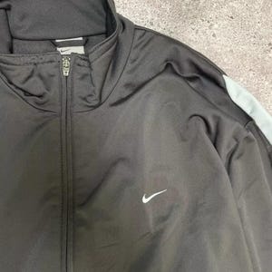 Puede incluir: Chaqueta deportiva Nike negra con cremallera completa y logotipo blanco de Nike. La chaqueta tiene una franja azul claro en el hombro y un cuello alto.