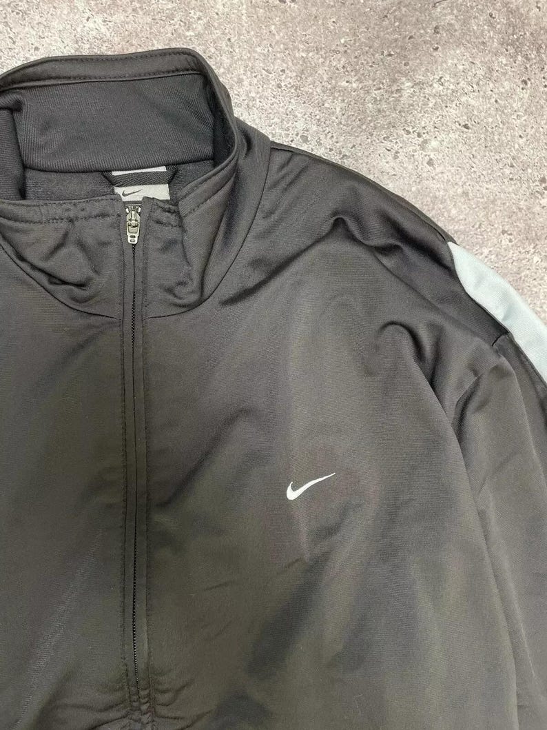 Puede incluir: Chaqueta deportiva Nike negra con cremallera completa y logotipo blanco de Nike. La chaqueta tiene una franja azul claro en el hombro y un cuello alto.