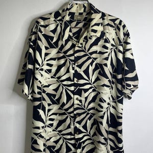 Puede incluir: Camisa de manga corta con un estampado de hojas en negro y crema. La camisa está colgada de una percha metálica sobre un fondo blanco. La camisa tiene cuello y un corte holgado.