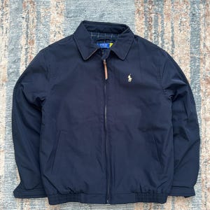 Könnte beinhalten: Marineblaue Jacke mit Reißverschluss, Kragen und braunem Leder-Reißverschluss. Das Polo by Ralph Lauren Logo in Gold ist auf der Brust zu sehen. Die Jacke hat einen gerippten Saum und Bündchen.