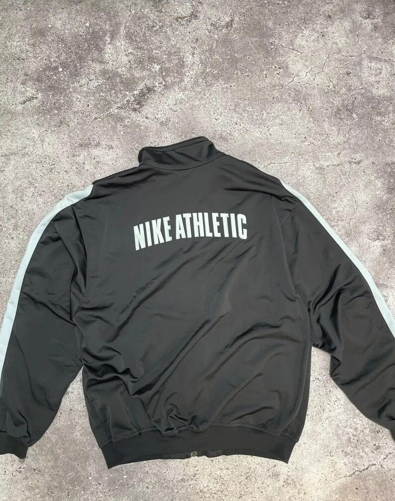 Puede incluir: Chaqueta deportiva negra con la inscripci&oacute;n "NIKE ATHLETIC" en blanco en la espalda. La chaqueta tiene un cuello alto y rayas azul claro en las mangas. La chaqueta es de un material brillante.
