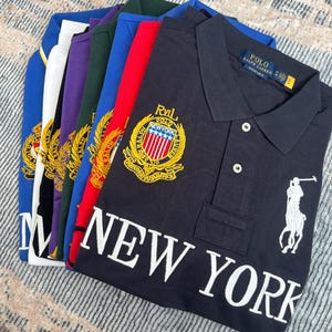 May include: A stack of polo shirts in various colors, including navy, red, and blue. The navy shirt features a white embroidered logo and the words "NEW YORK". Each shirt has a collared neckline and button closure.