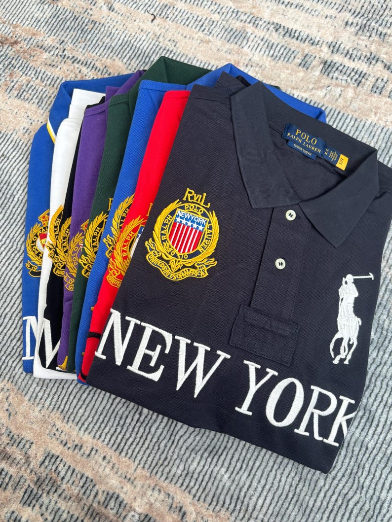 May include: A stack of polo shirts in various colors, including navy, red, and blue. The navy shirt features a white embroidered logo and the words "NEW YORK". Each shirt has a collared neckline and button closure.
