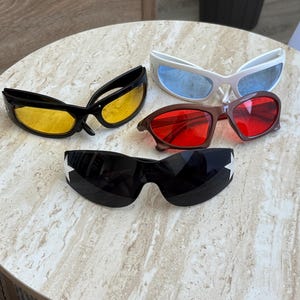 May include: Four pairs of wraparound sunglasses with distinct frames and lenses. One pair has black frames with yellow lenses, one white with blue lenses, one brown with red lenses, and one black with dark lenses featuring white star accents on the sides.