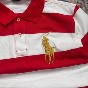 Puede incluir: Polo a rayas rojas y blancas con cuello cl&aacute;sico y tapeta abotonada. Presenta un logotipo dorado bordado de un jugador de polo a caballo. La camisa tiene el texto "POLO RALPH LAUREN" en la etiqueta.