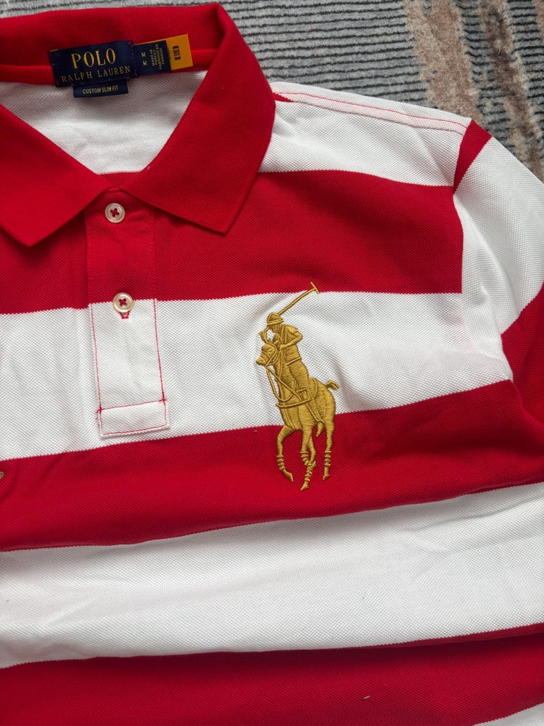 Puede incluir: Polo a rayas rojas y blancas con cuello cl&aacute;sico y tapeta abotonada. Presenta un logotipo dorado bordado de un jugador de polo a caballo. La camisa tiene el texto "POLO RALPH LAUREN" en la etiqueta.
