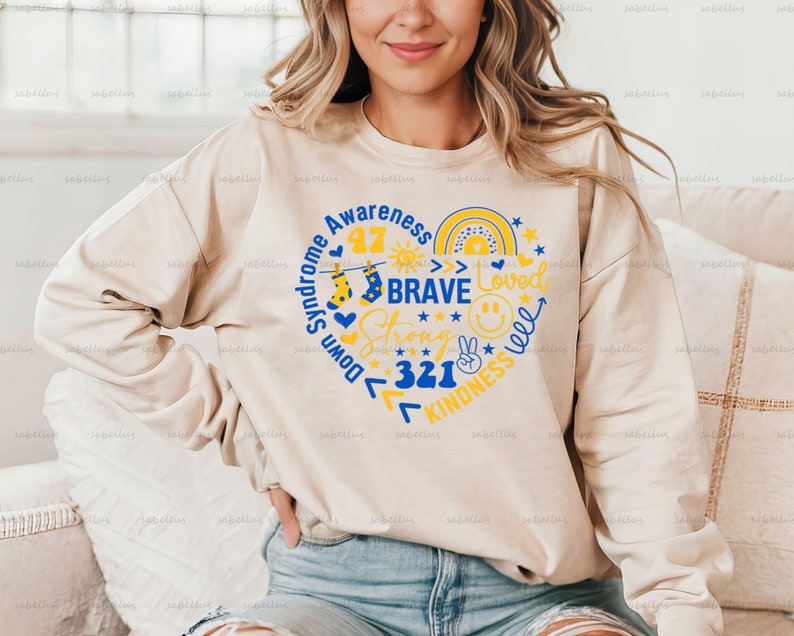 May include: A beige sweatshirt with a blue and yellow heart design. The heart includes the words "Down Syndrome Awareness", "Brave", "Strong", "Loved", "Kindness", and "321".