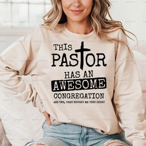 May include: A beige sweatshirt with black text that reads "This Pastor Has An Awesome Congregation And Yes, They Bought Me This Shirt". A cross is above the word "Pastor".