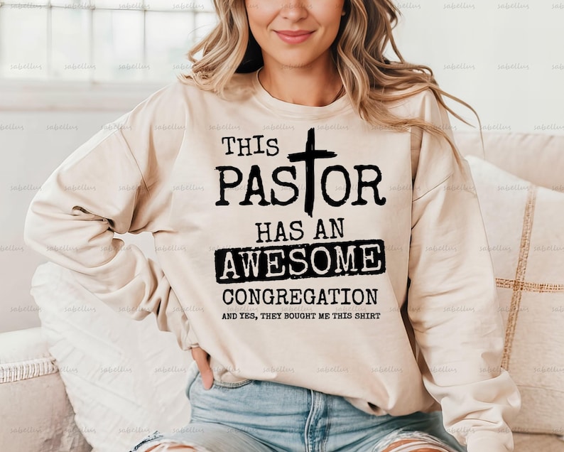 May include: A beige sweatshirt with black text that reads "This Pastor Has An Awesome Congregation And Yes, They Bought Me This Shirt". A cross is above the word "Pastor".