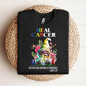 May include: Black t-shirt with a colorful graphic of a gnome wearing a rainbow hat and holding a ribbon. The text "HEAL CANCER" is above the gnome and the text "FOR WITH GOD NOTHING IS IMPOSSIBLE LUKE 1:37" is below the gnome.