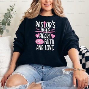 May include: A black sweatshirt with the text "Pastor's Wife Heart Full of Faith and Love" in pink lettering. The sweatshirt has a pink heart with a cross in the center.