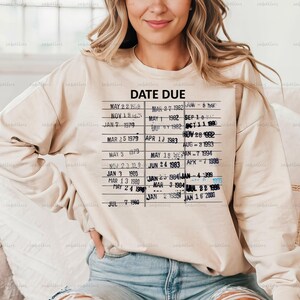 Pu&ograve; includere: Una felpa beige con un design di carta di biblioteca vintage che presenta il testo "DATE DUE" e un elenco di date da maggio 1979 a gennaio 2000.