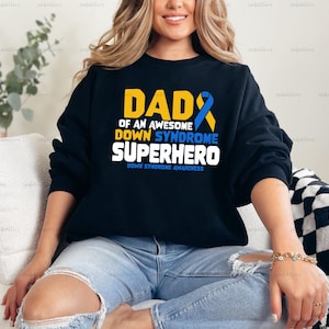 May include: A black sweatshirt with the text "Dad of an Awesome Down Syndrome Superhero" in yellow and blue. The text "Down Syndrome Awareness" is printed in smaller font below the main text.