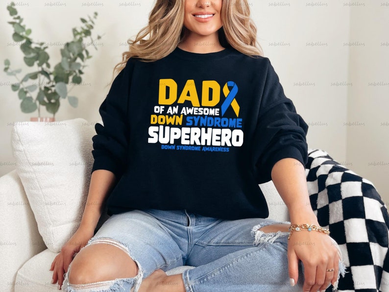 May include: A black sweatshirt with the text "Dad of an Awesome Down Syndrome Superhero" in yellow and blue. The text "Down Syndrome Awareness" is printed in smaller font below the main text.