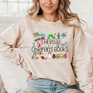 Pu&ograve; includere: Una felpa beige con una stampa grafica colorata di personaggi di libri per bambini e il testo "I Still Read Children's Books".