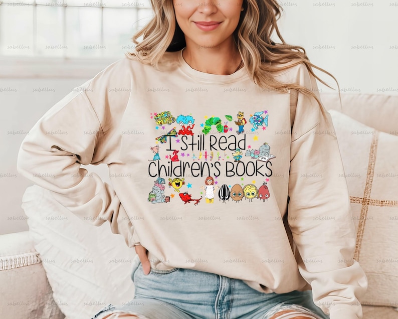 Pu&ograve; includere: Una felpa beige con una stampa grafica colorata di personaggi di libri per bambini e il testo "I Still Read Children's Books".