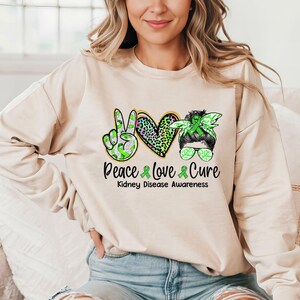 May include: A light beige sweatshirt with a green ribbon peace sign, a green ribbon heart with a leopard print, and a green ribbon headband with sunglasses. The text "Peace Love Cure" is printed below the design, with the words "Kidney Disease Awareness" printed below the text.