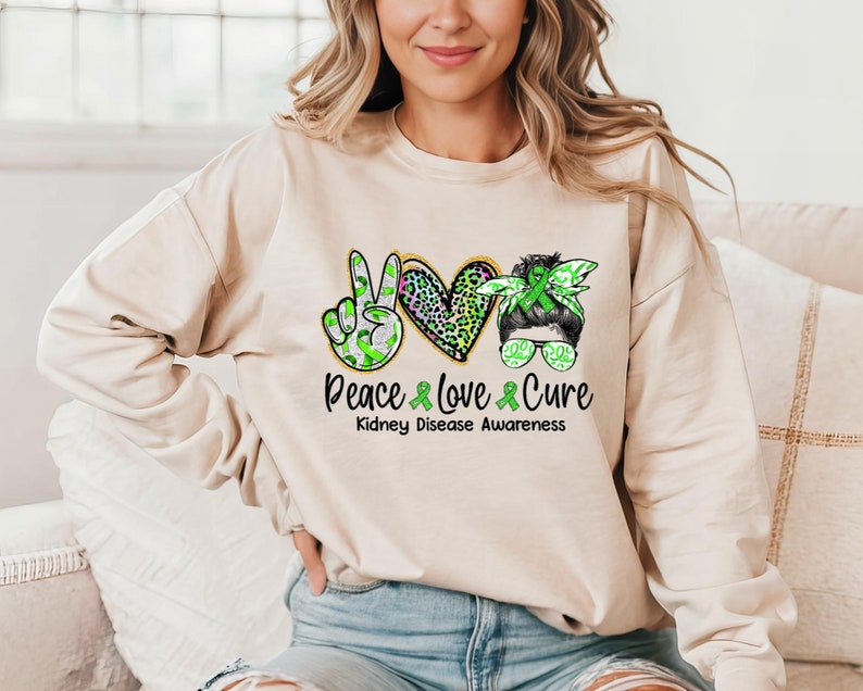 May include: A light beige sweatshirt with a green ribbon peace sign, a green ribbon heart with a leopard print, and a green ribbon headband with sunglasses. The text "Peace Love Cure" is printed below the design, with the words "Kidney Disease Awareness" printed below the text.