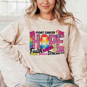 May include: A beige sweatshirt with a pink ribbon and the words "Fight Cancer" and "Hope" in colorful letters. The words "Faith", "Brave", "Love", "Power", "Believe", and "Strength" are also included in the design.