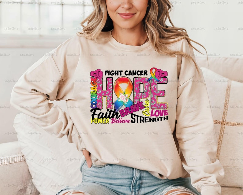May include: A beige sweatshirt with a pink ribbon and the words "Fight Cancer" and "Hope" in colorful letters. The words "Faith", "Brave", "Love", "Power", "Believe", and "Strength" are also included in the design.