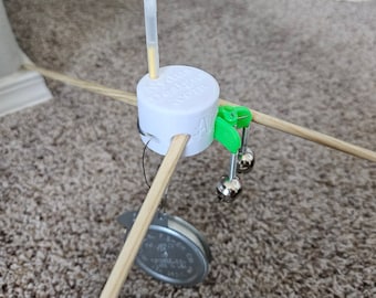 May include: A white plastic mobile holder with a yellow dropper attached. The holder has four wooden dowels attached to it, and a silver bell and a silver lid are hanging from the dowels.