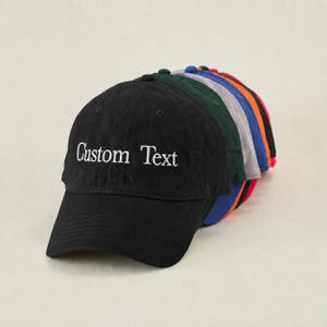 Puede incluir: Una pila de gorras de béisbol en varios colores, incluyendo negro, verde, gris, azul, naranja y rojo. La gorra negra delantera tiene el texto bordado "Custom Text" en blanco.