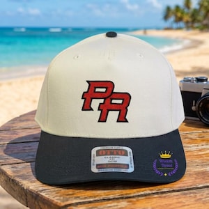 May include: A two-tone baseball cap with a cream-colored crown and a black brim. The front features the letters PR embroidered in red with a black outline. A sticker on the brim reads OTTO Classic Fit, and a small logo with a crown is visible.