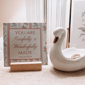 May include: A white ceramic swan ring holder with a gold accent on the beak. The swan is sitting on a book with the title "Once Upon A Time" printed in gold lettering. A small card with the text "You Are Fearfully & Wonderfully Made Psalm 139:14" is standing upright on a wooden stand.